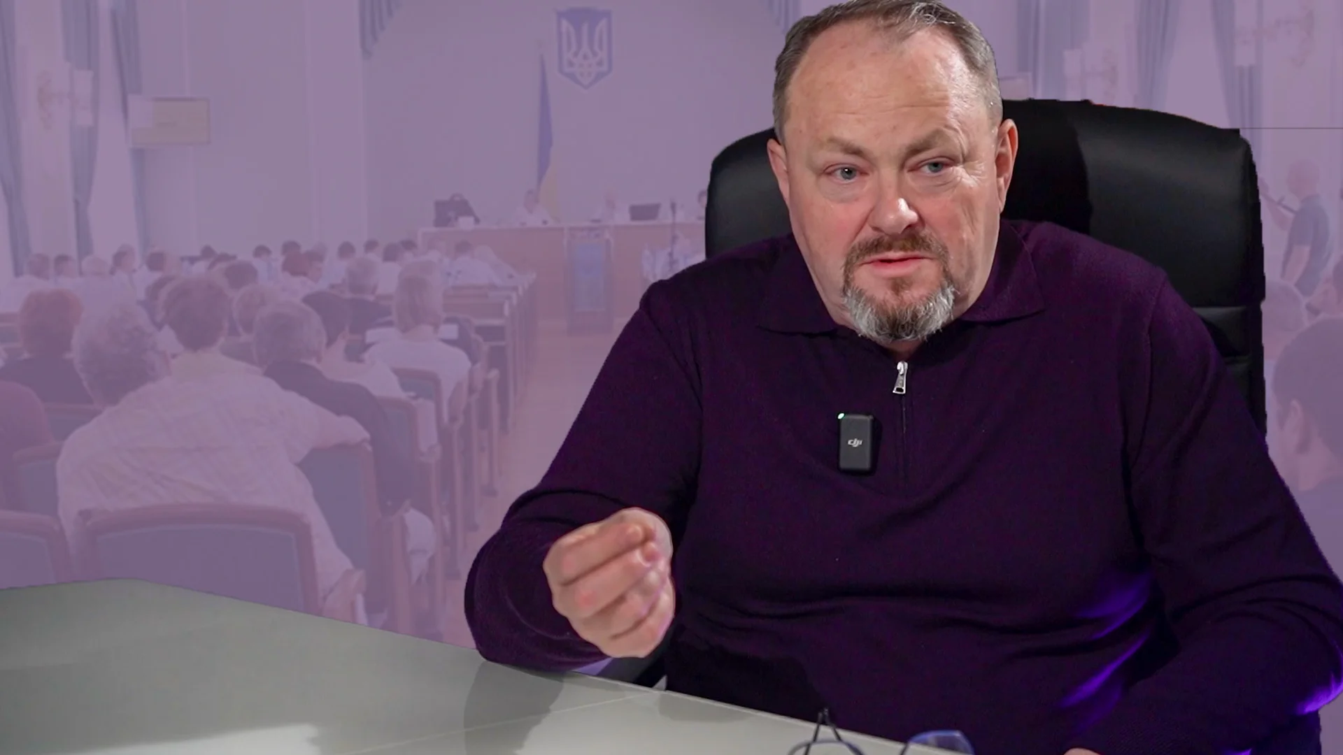 Man in a purple sweater sits at a desk with a clip-on mic, gesturing while speaking, audience blurred in the background.